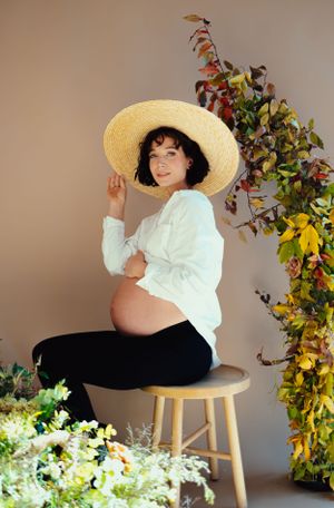 A maternity photo featuring a woman in white shirt and straw hat posing on a stool with floral decorations.