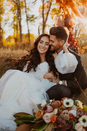 Intimate autumn wedding photo with couple sitting in golden leaves with colorful floral bouquet.