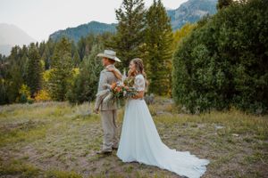 Wedding couple stands in mountain meadow wearing rustic attire.