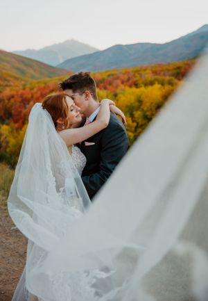 Wedding couple embracing with mountain fall foliage and flowing veil in background.