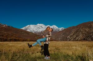 A playful couple lifts each other in a mountain meadow with snowy peaks in background.
