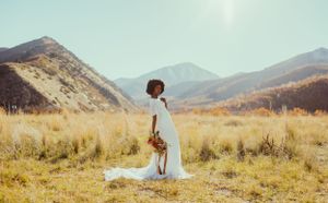 Bride in white dress stands in scenic mountain meadow with wildflowers and distant peaks.