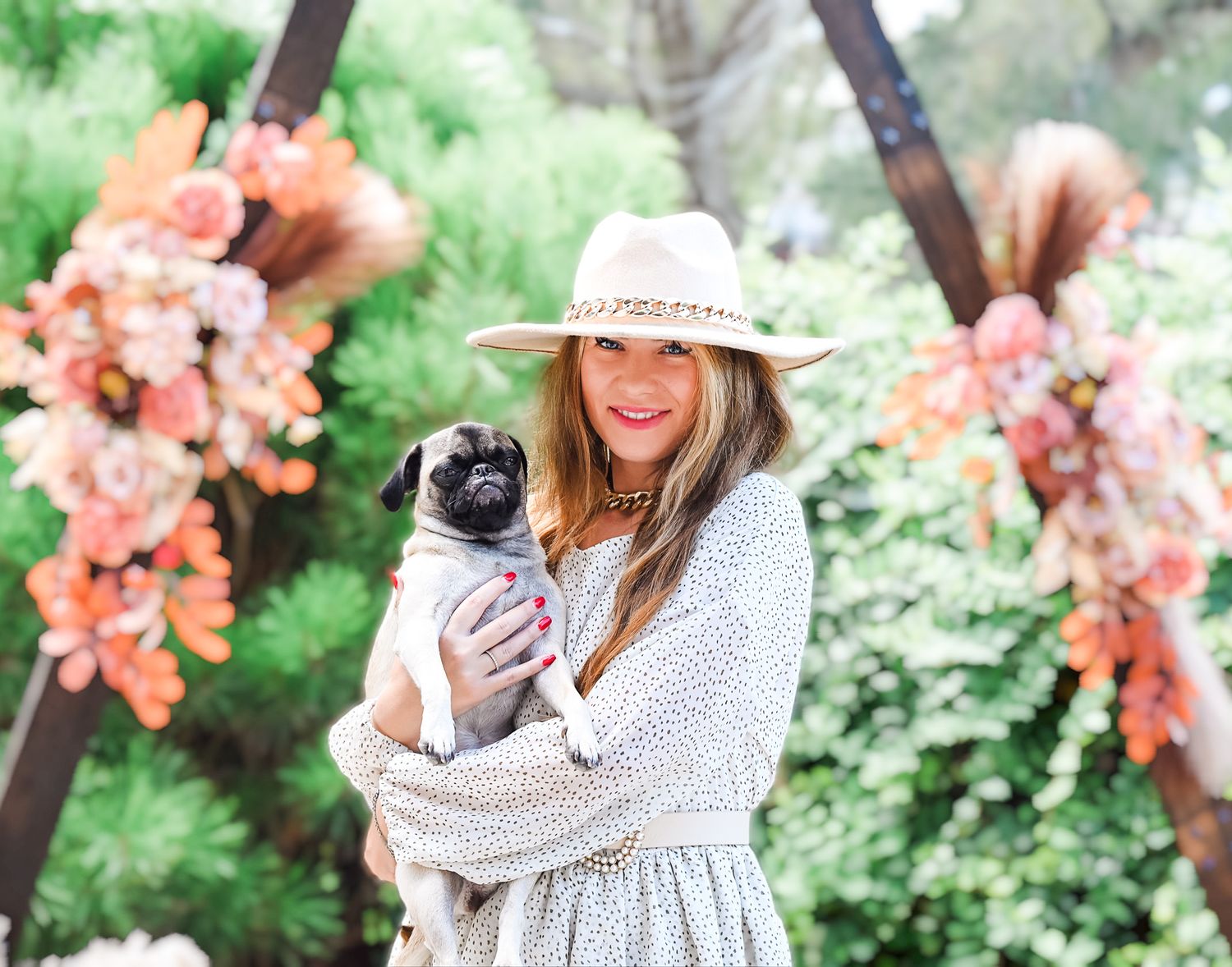 Person in white dress and straw hat holding small dog near floral arch with coral blooms.