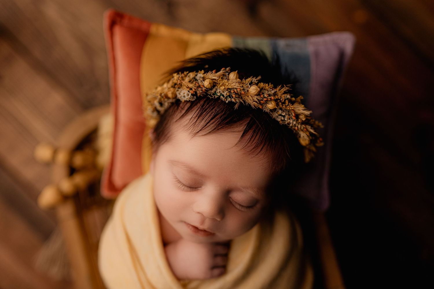 Sleeping newborn with delicate floral crown wrapped in cream swaddle against colorful pillows.