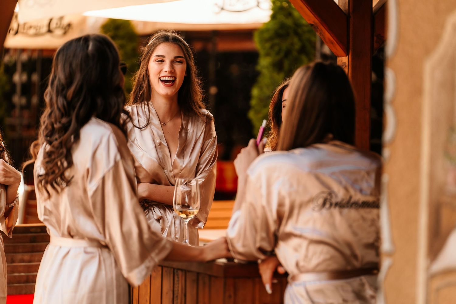 Friends in silk robes sharing drinks and laughs on a rustic wooden porch decorated with flowers.