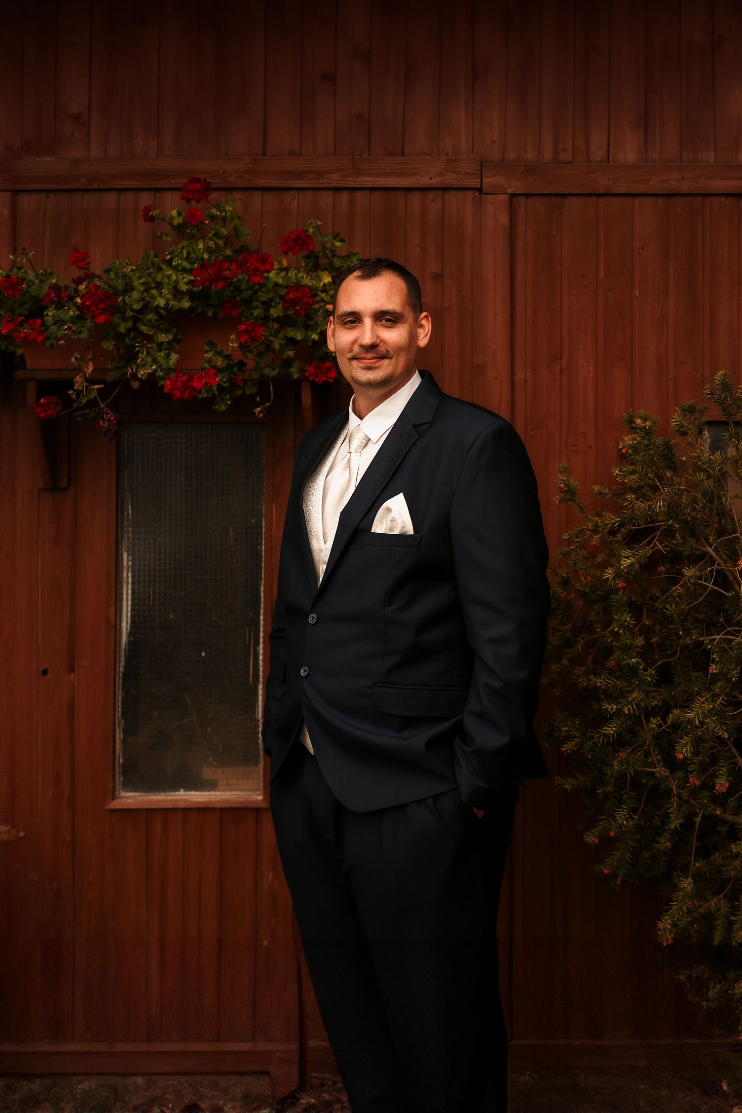 A groom in a classic black tuxedo with white vest poses against a rustic brown wall with red flowers.