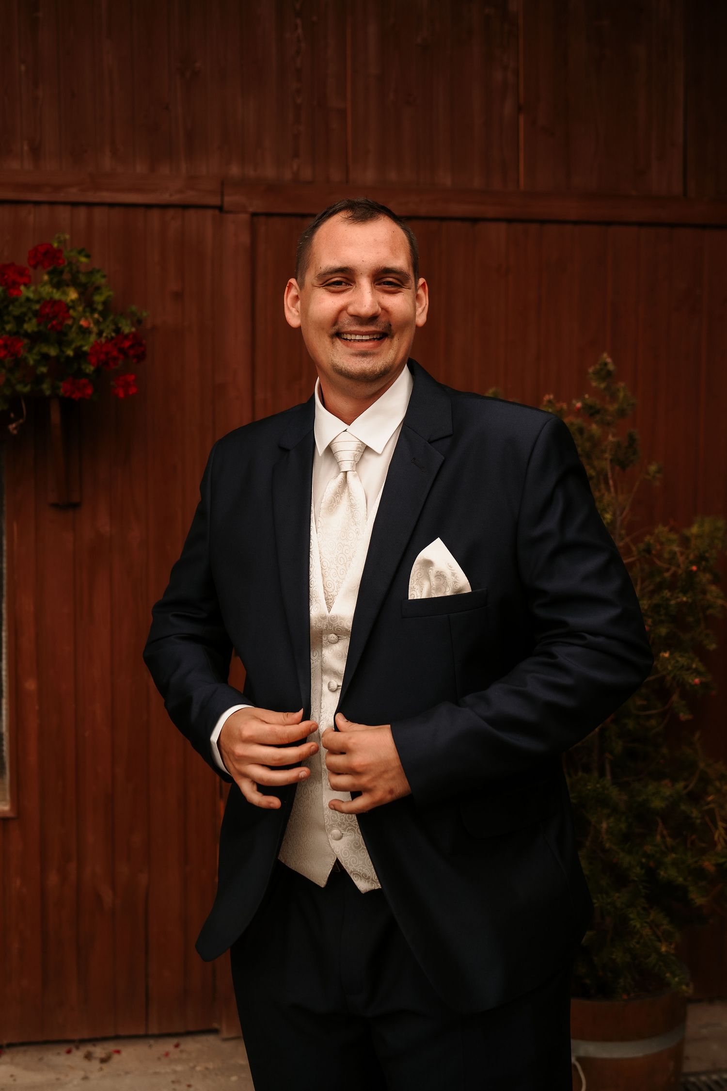 Wedding portrait showing formal attire and white pocket square against wooden backdrop with red flowers.