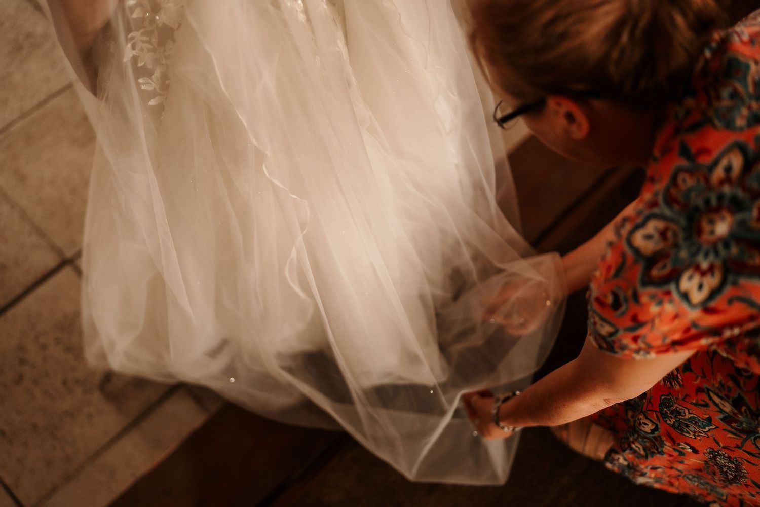 Hands adjust the layers of a flowing white wedding dress against patterned flooring.