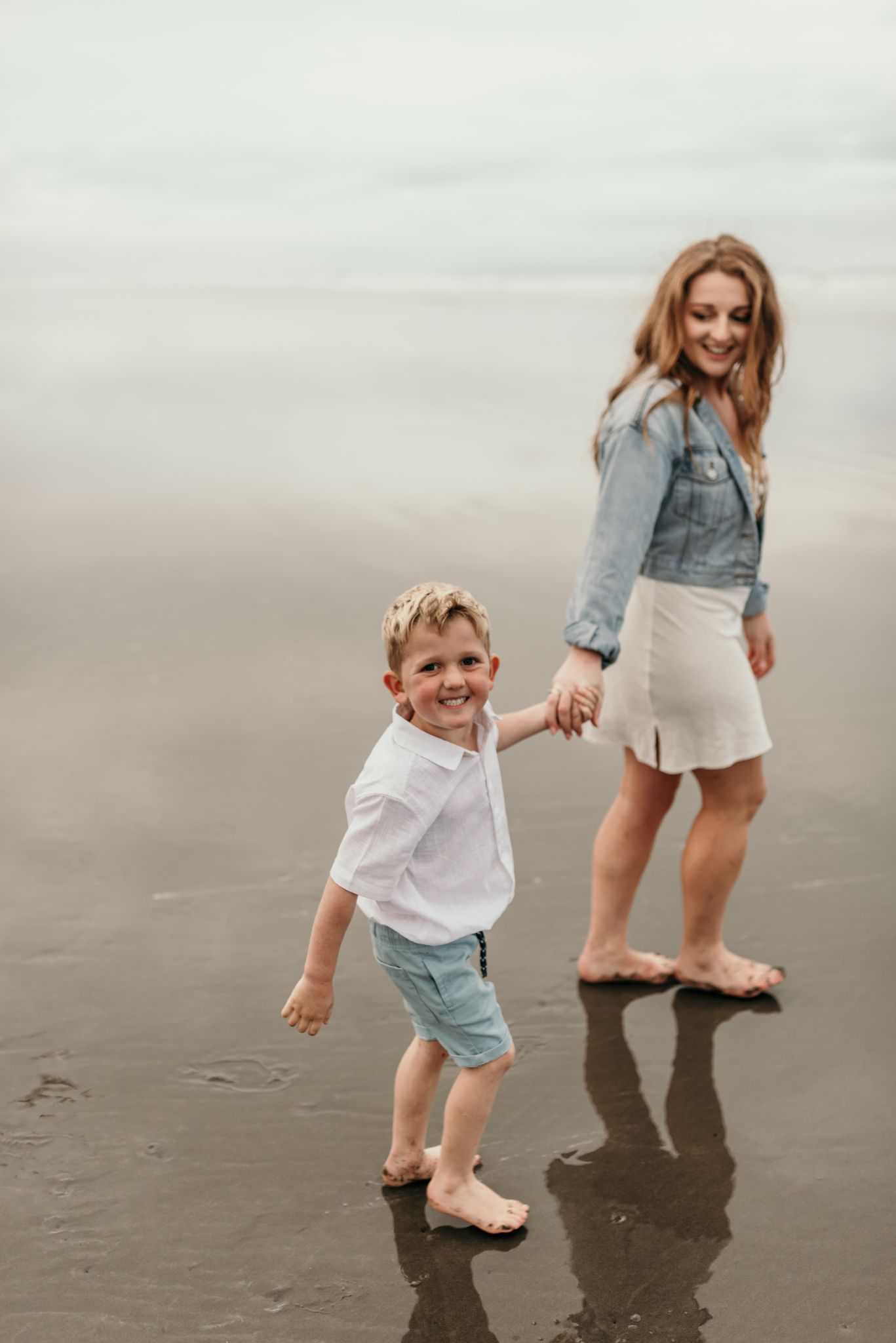 A joyful barefoot moment on the beach as two people hold hands and smile against an overcast sky.