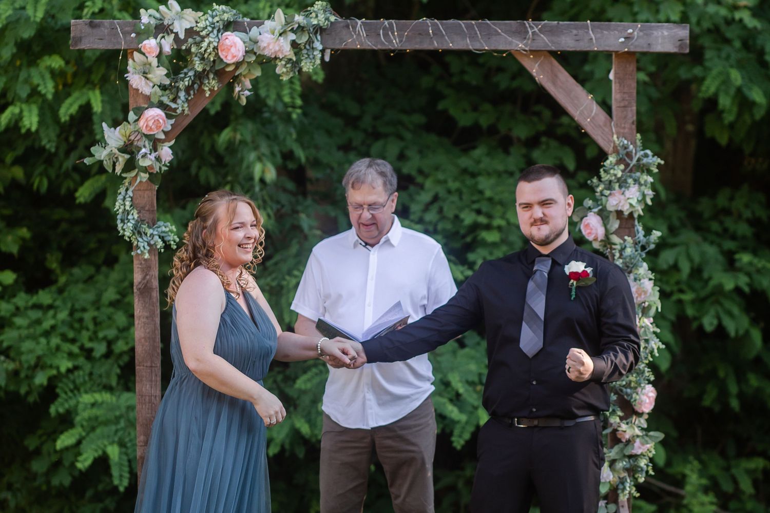 Wedding ceremony participants standing beneath a rustic wooden arch adorned with floral arrangements.