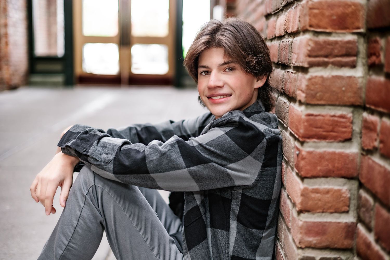 Indoor portrait wearing plaid shirt while sitting against brick wall.
