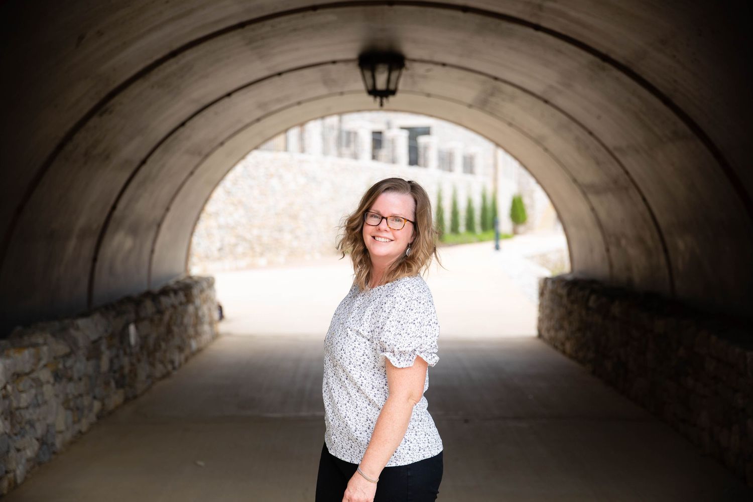 Person standing under curved stone tunnel in casual attire.