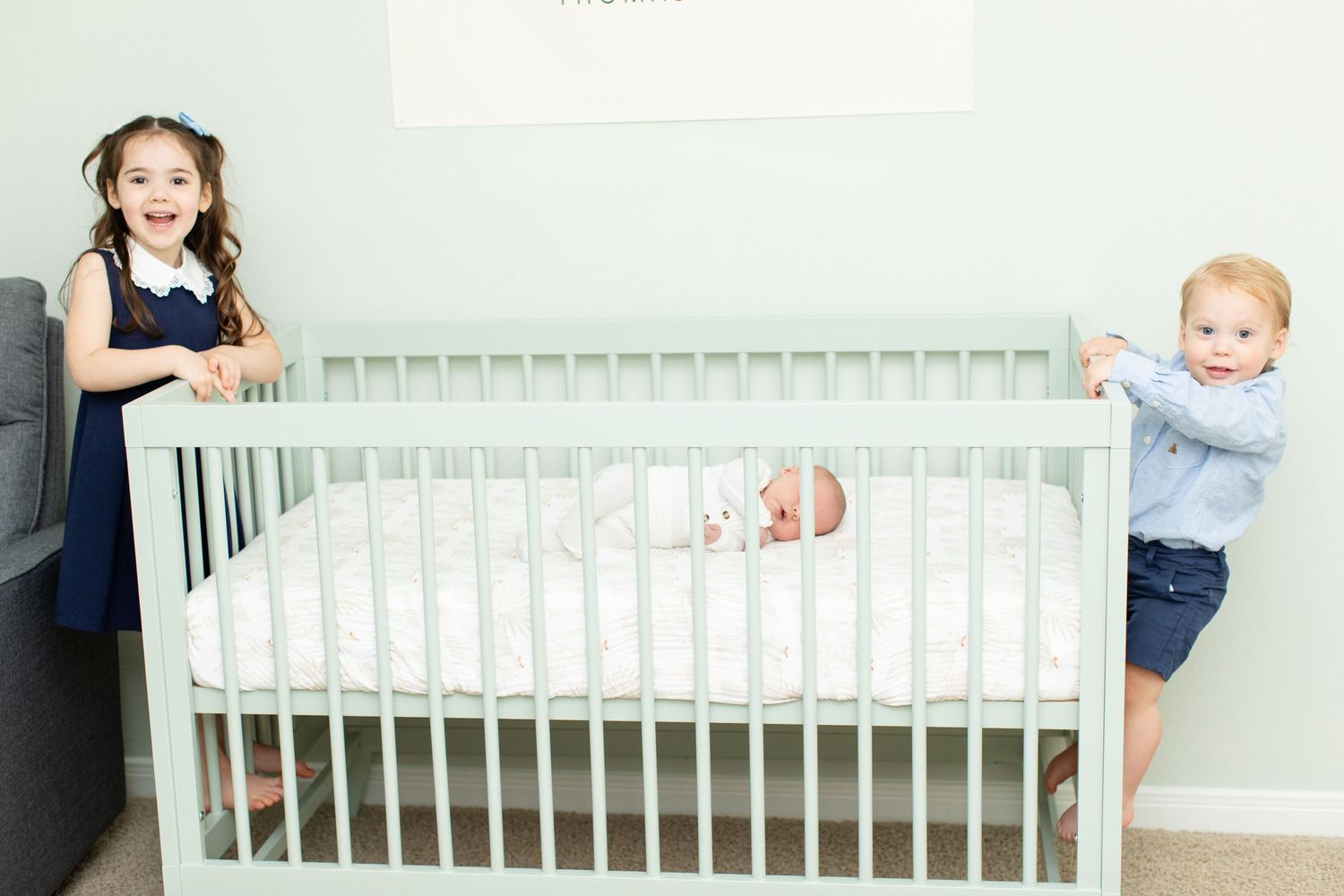 Children peek over sides of mint green crib at baby sibling in nursery setting.