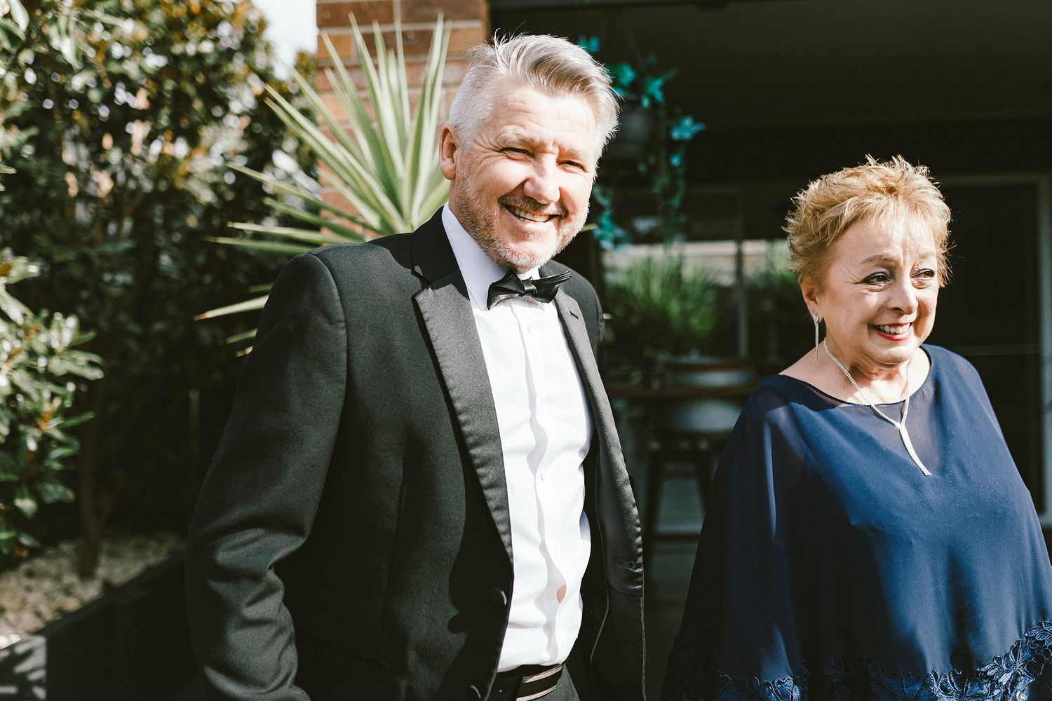 A couple in formal attire share a happy moment in front of a garden setting.