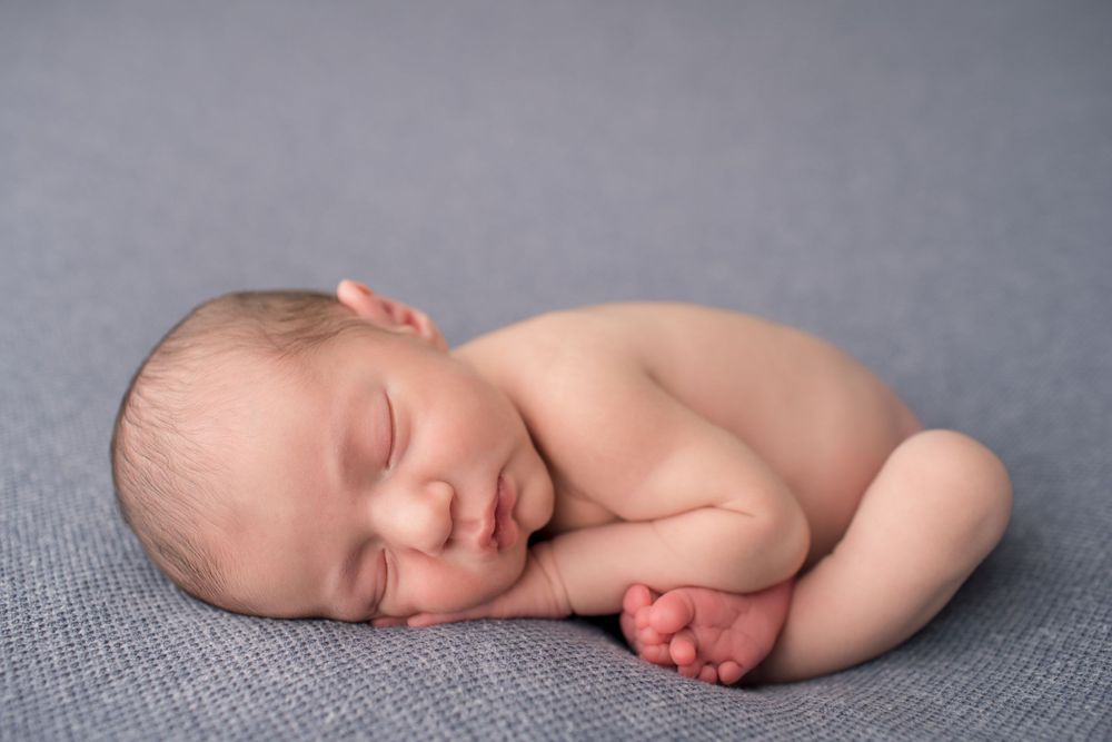 Side view of sleeping newborn baby in fetal position on gray fabric.
