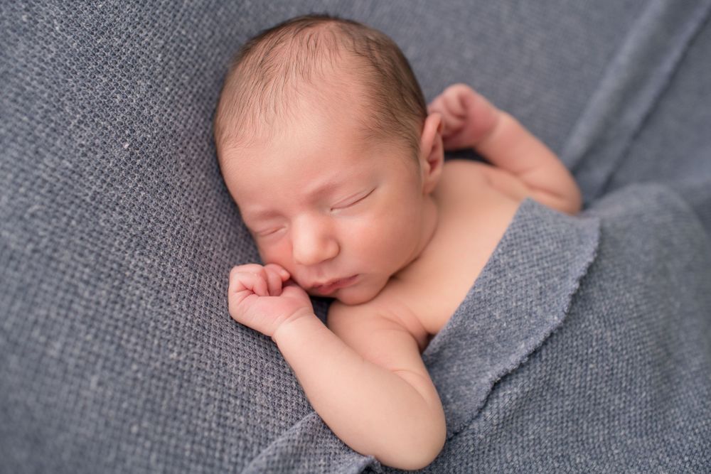Newborn baby sleeping peacefully on gray fabric with hands near face.