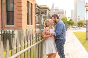 Romantic couple embracing by historic brick building in warm evening light with city skyline in background.