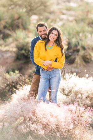 A couple poses together in a desert landscape with soft pink wildflowers in the foreground.