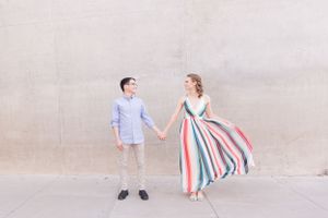 Couple poses playfully against neutral wall, with striped dress creating movement in the frame.