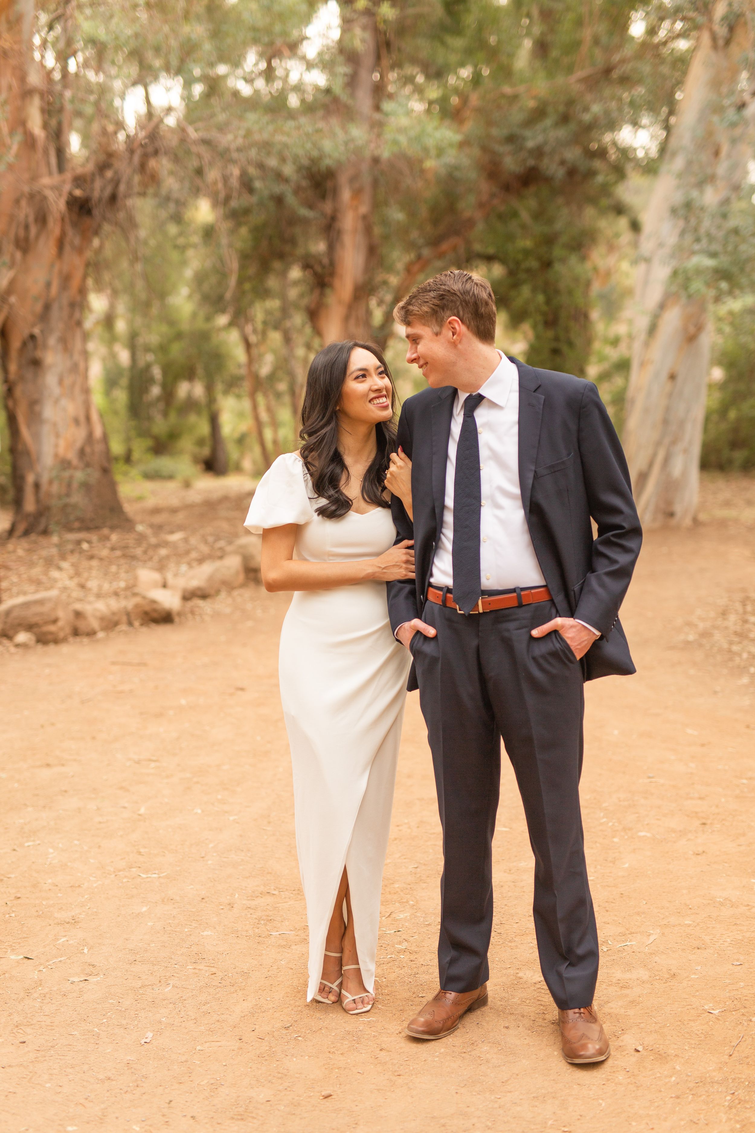 A couple shares an intimate moment on a dirt path surrounded by eucalyptus trees.