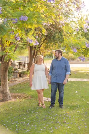 A couple strolls through a park with blooming jacaranda trees and scattered purple petals on the grass.
