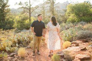 A couple walks hand in hand on a desert trail surrounded by cacti and mountains at golden hour.