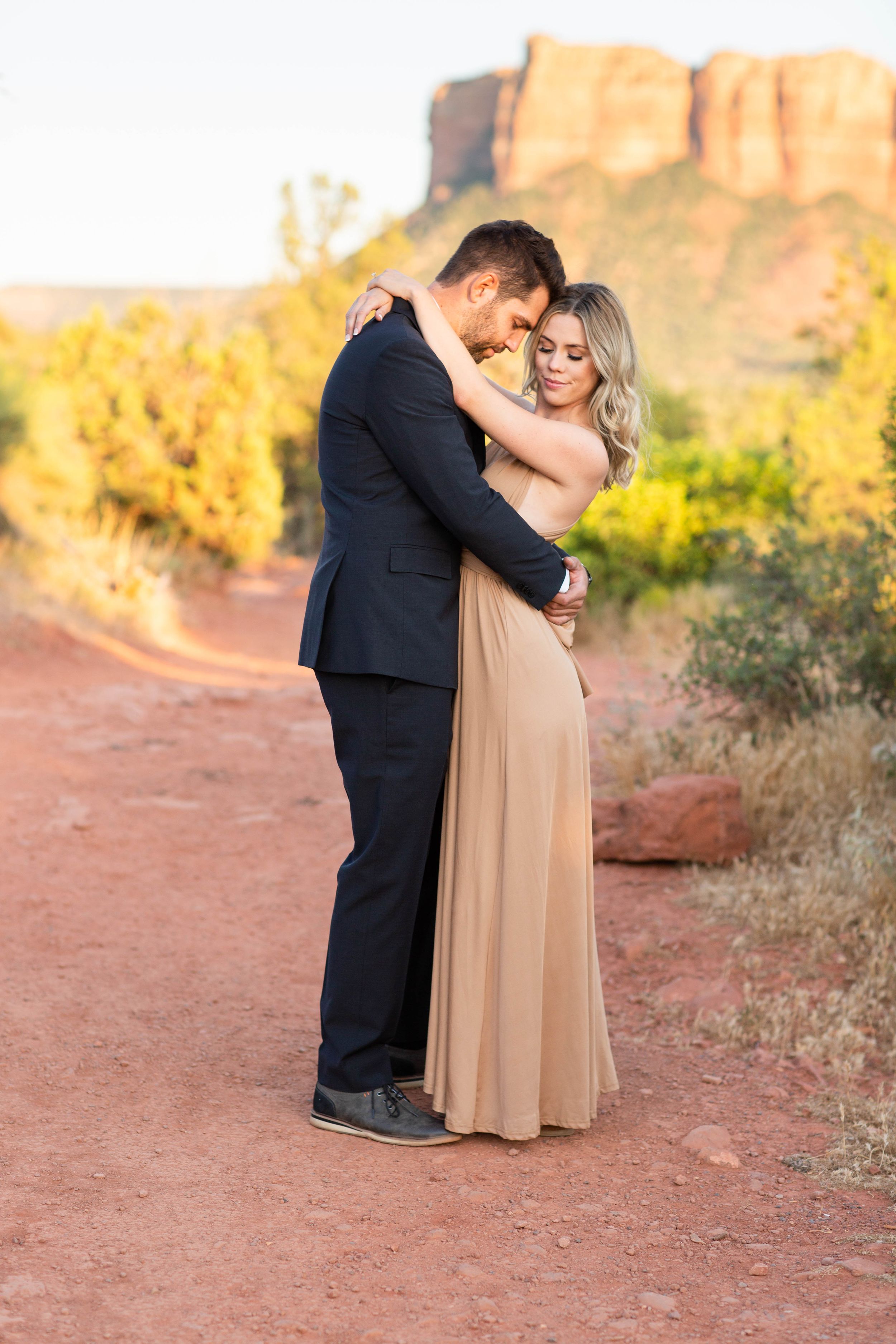 A romantic couple embraces on a red dirt path with dramatic desert cliffs and golden sunset lighting in the background.