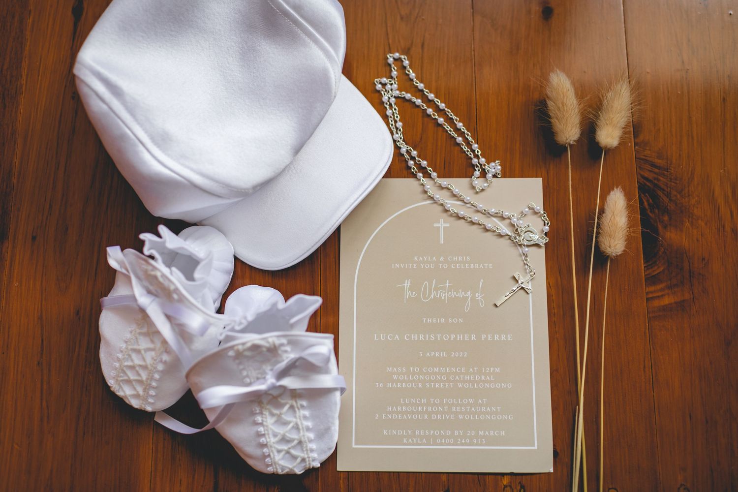 White baby baptism items including a cap, booties, and invitation card displayed with dried flowers on wooden surface.