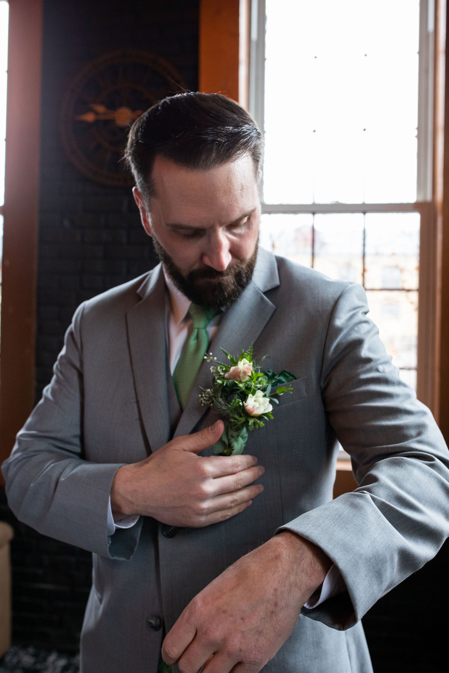 Groom in gray suit adjusts boutonniere while looking down thoughtfully.