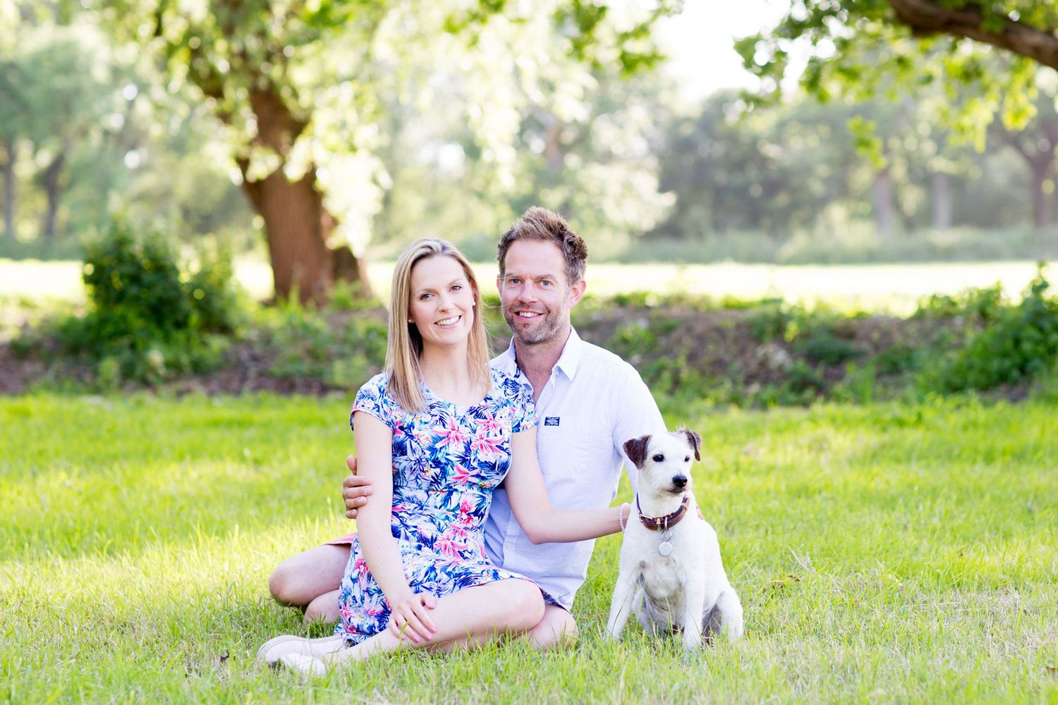 A couple sits on grass with their Jack Russell terrier dog during a sunny outdoor photoshoot.