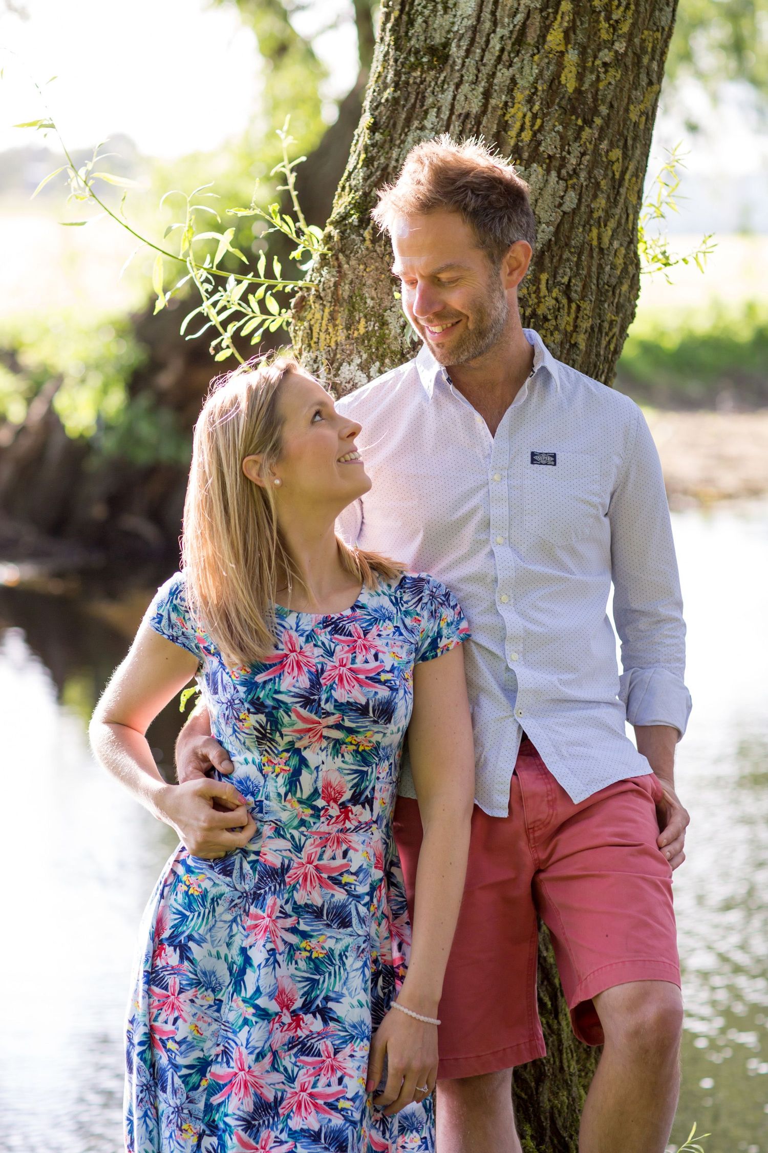 A couple shares an intimate moment leaning against a tree by the water.