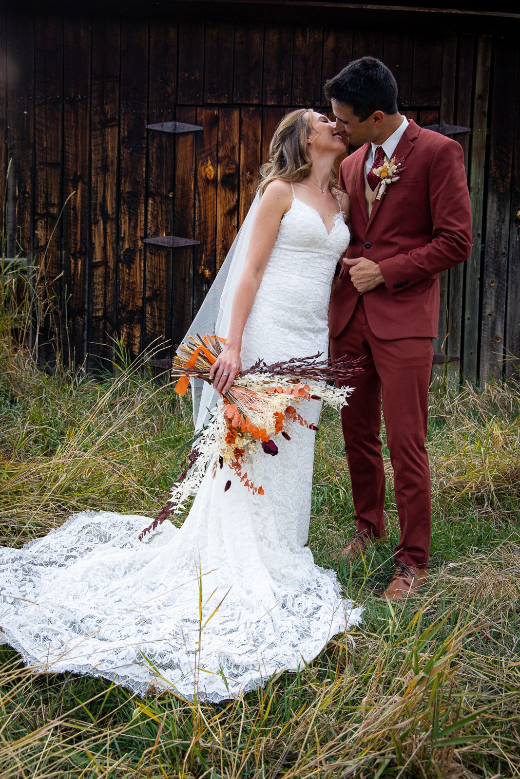 Rustic barn wedding portrait with bride holding autumn bouquet and burgundy suit details.
