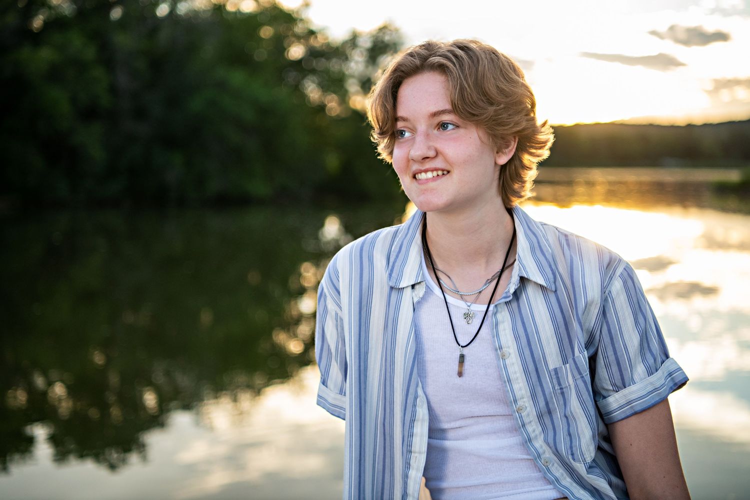 Candid sunset portrait by lake wearing striped button-down shirt and necklace.
