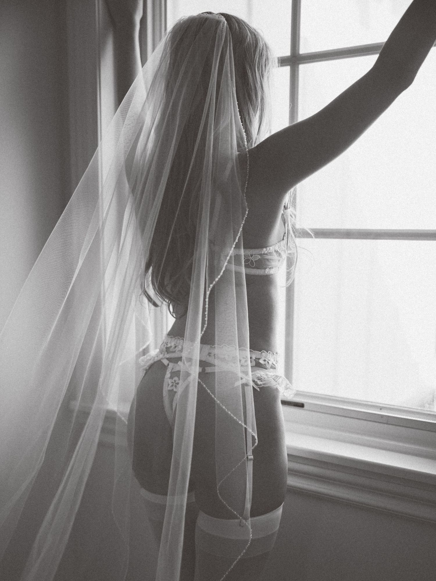 Silhouette of person in bridal lingerie and flowing veil by window in black and white.