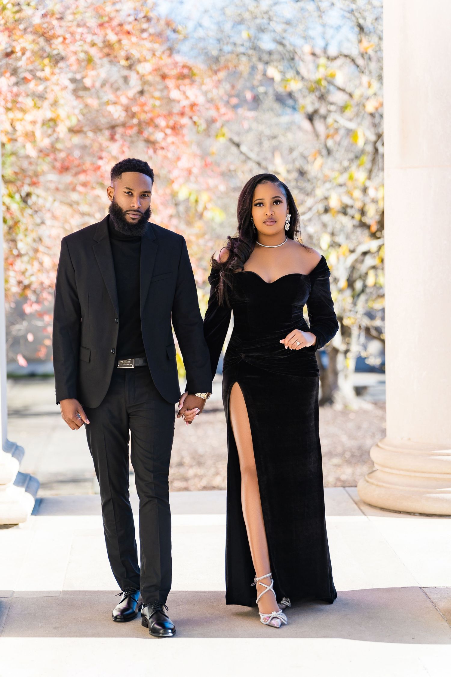 An elegant couple in formal black attire poses outdoors during autumn.