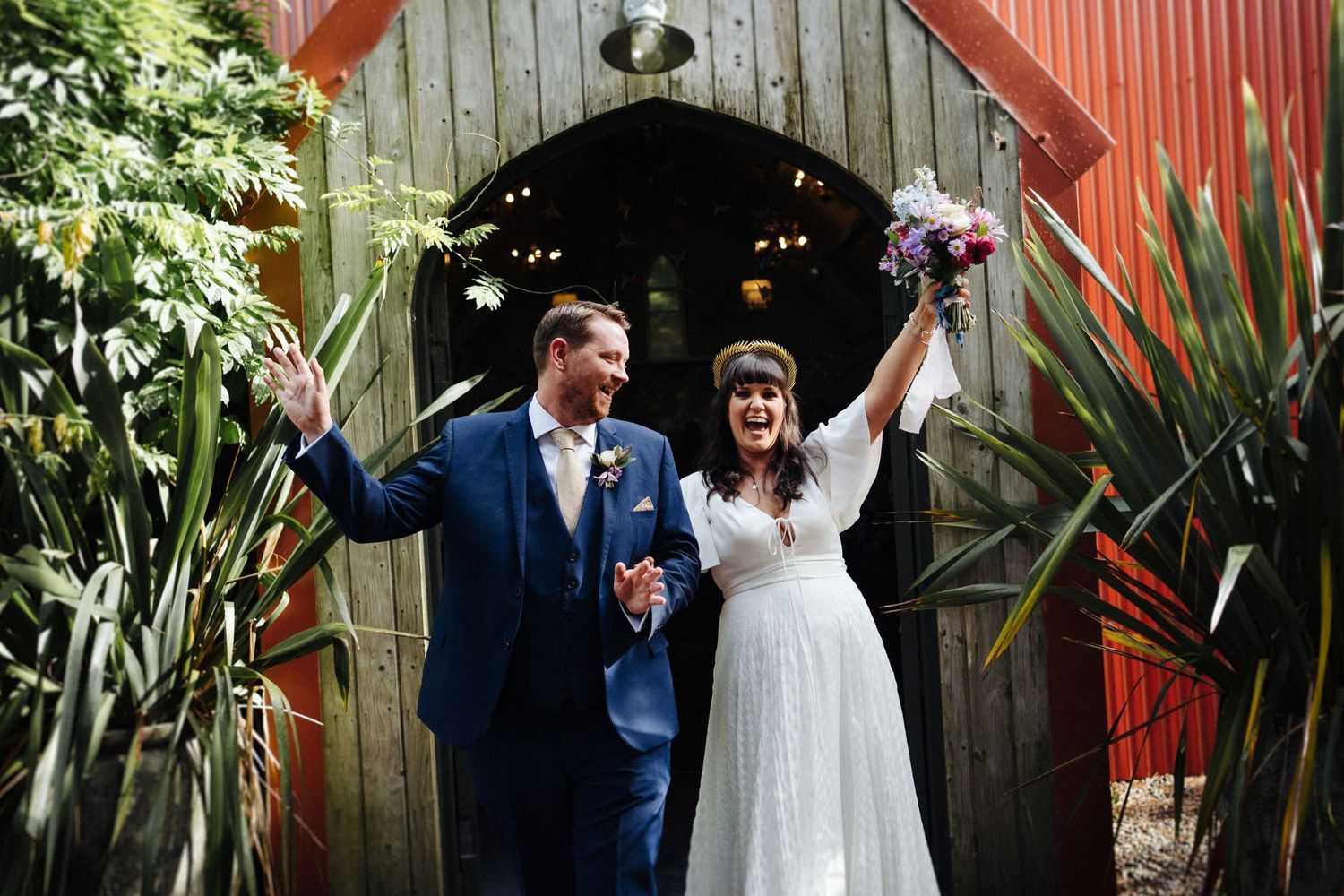 Newlyweds celebrating with raised arms in front of a rustic red barn entrance.