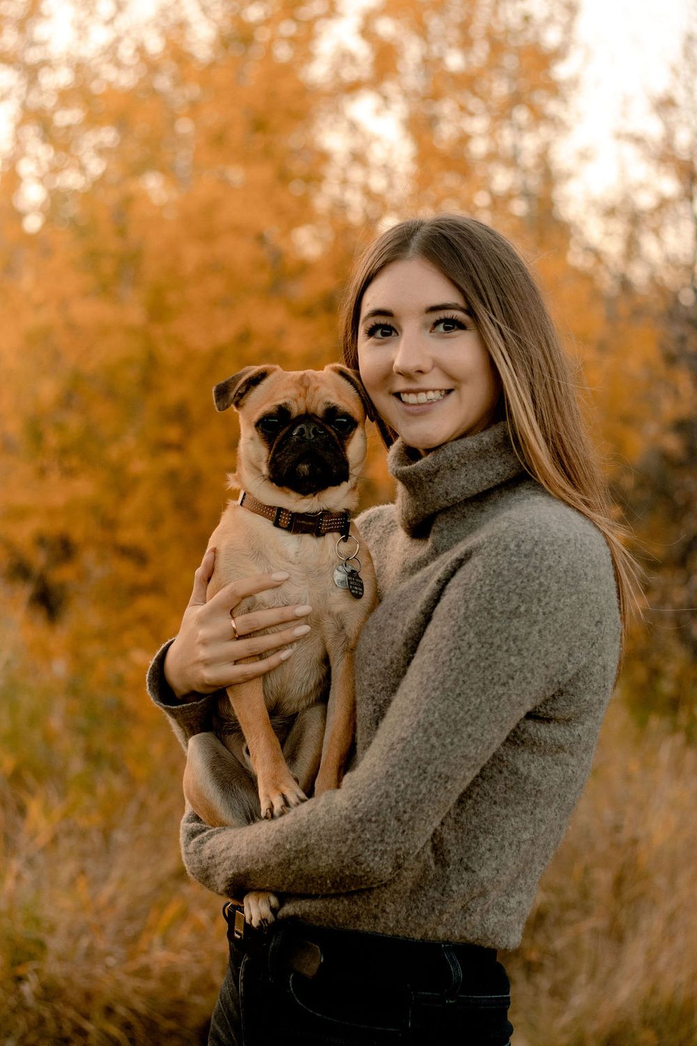Girl holding her pug in front of orange fall trees both looking at the camera and she is smiling.