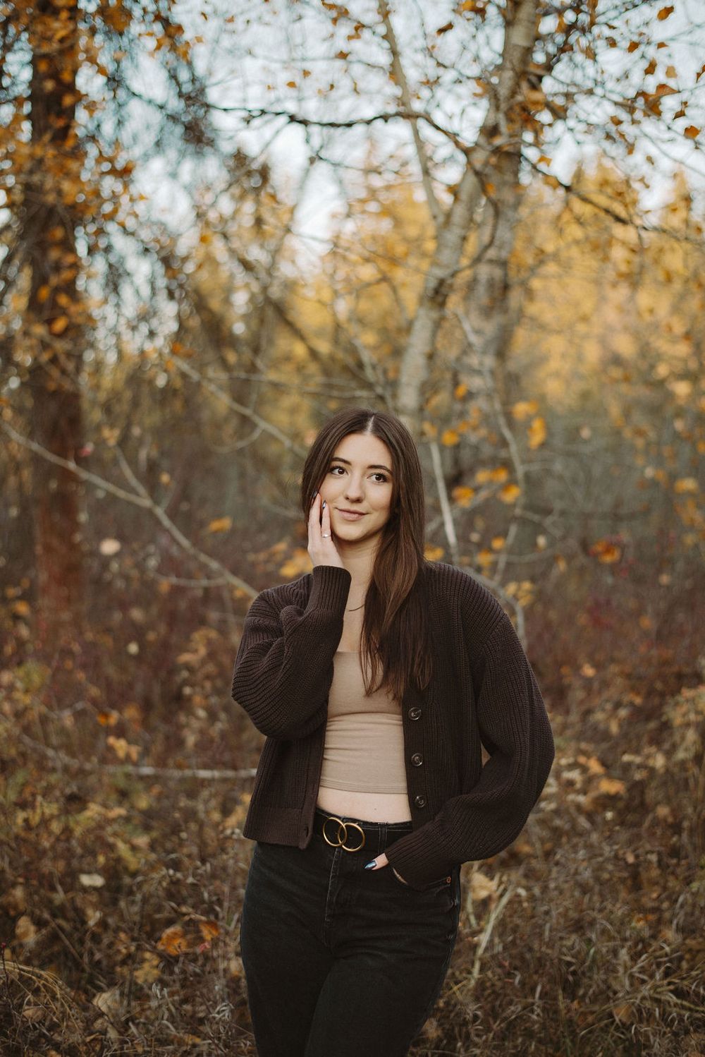Girl wearing brown standing in front of fall trees with one hand on her cheek.