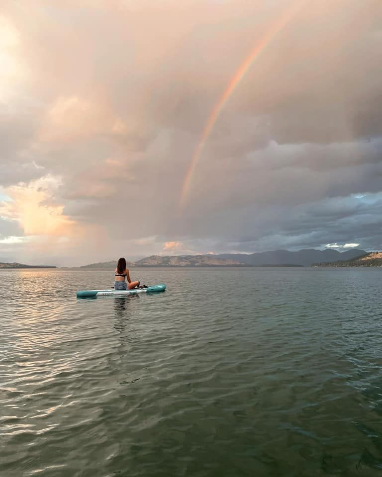 Girl on paddleboard on a lake with a rainbow in the sky.