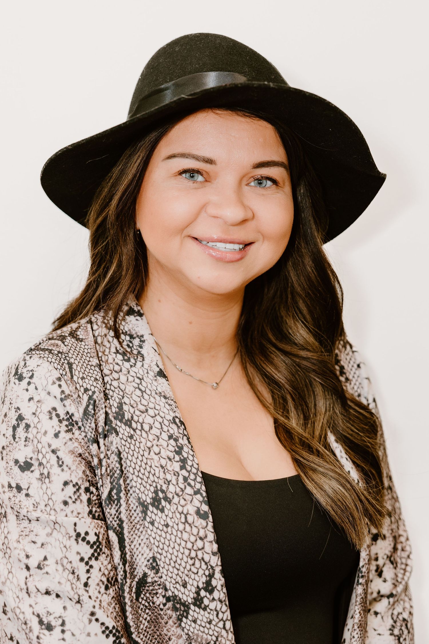 A person wearing a black hat and snake print cardigan smiles in a professional headshot.