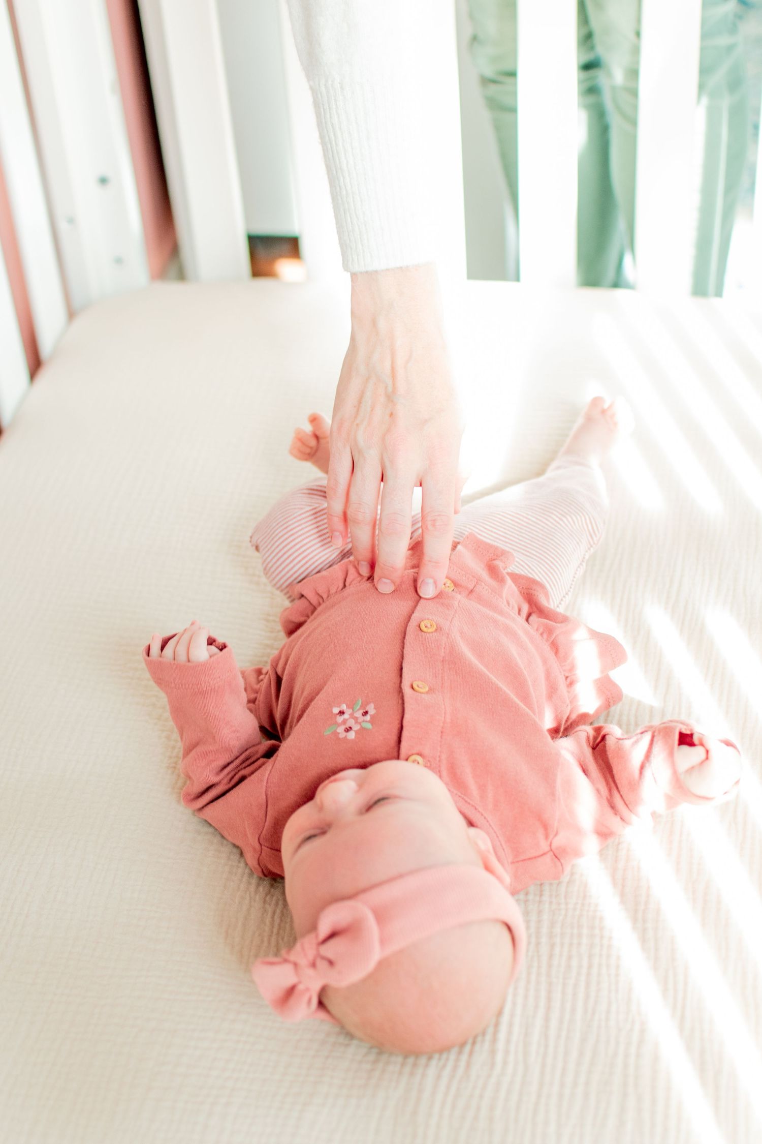 Infant wearing coral pink onesie lying on white crib mattress in natural light.
