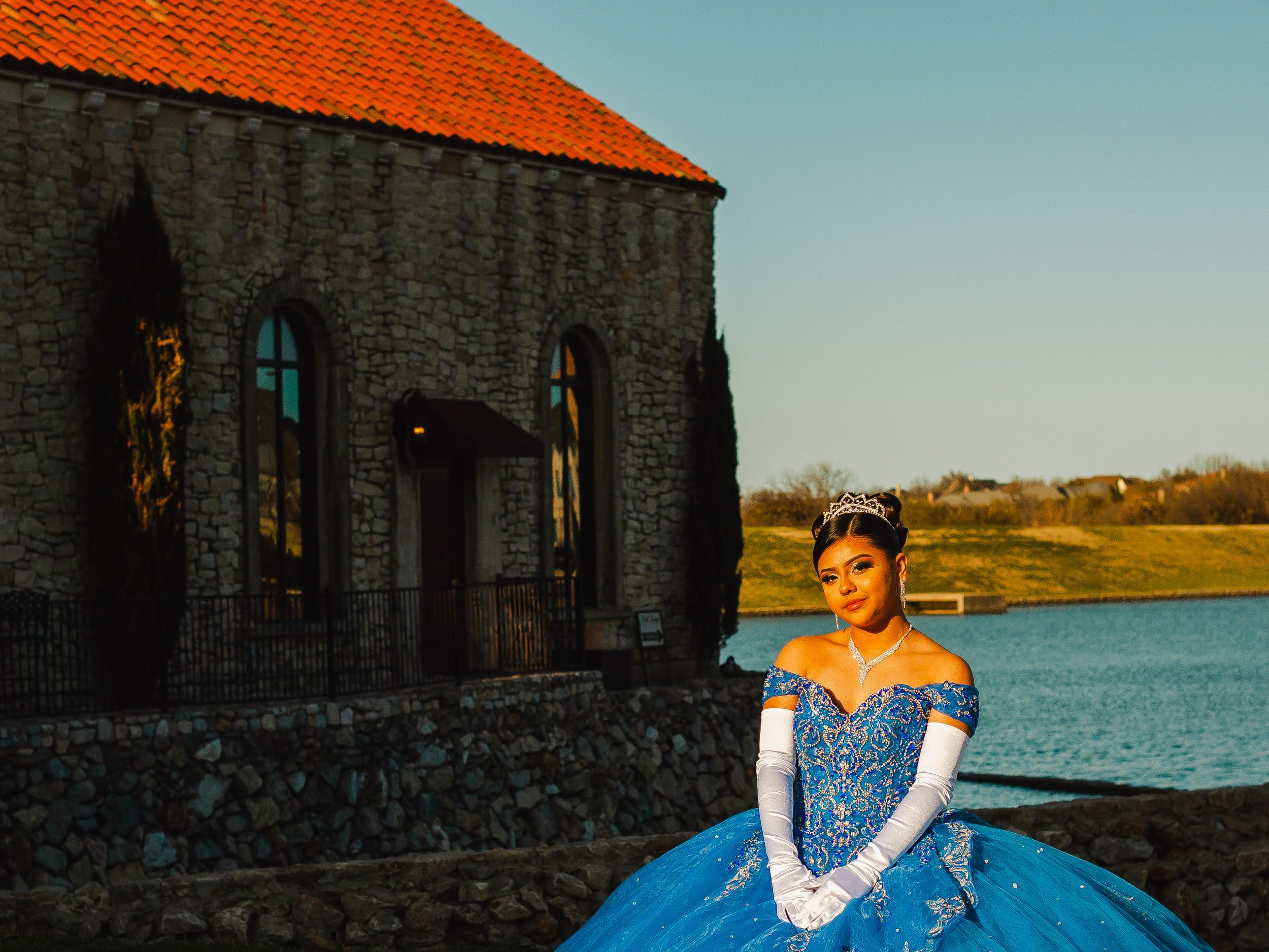 A blue formal dress photographed near a stone building with orange roof tiles and waterfront view.