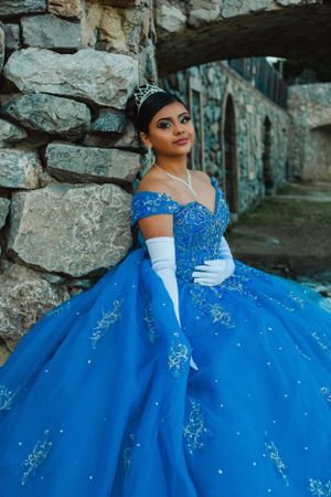 Sparkling blue formal dress with off-shoulder design photographed against stone wall.