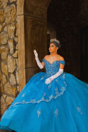 Elegant off-shoulder blue ball gown with white gloves against stone architecture.