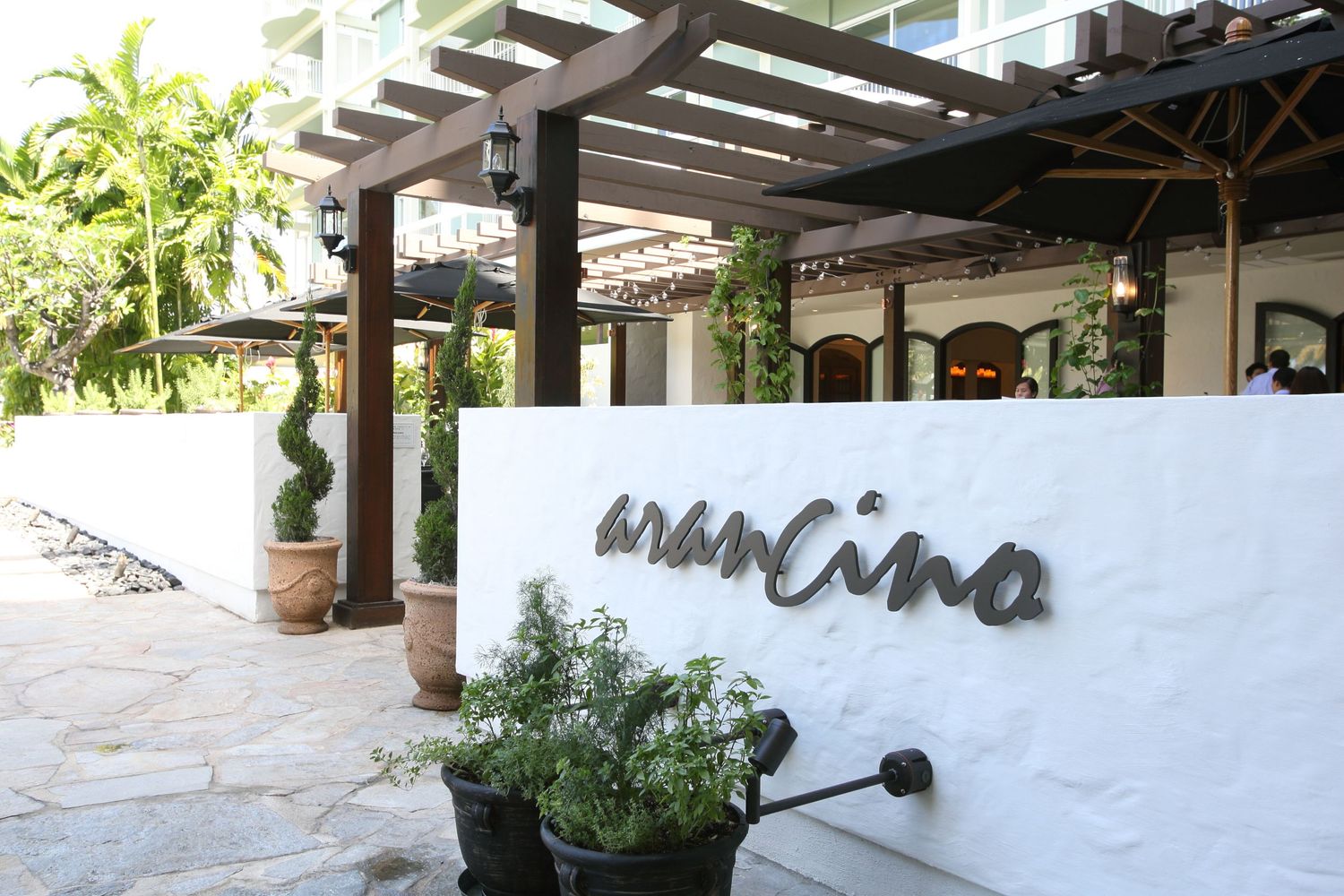 Exterior entrance of Mancino restaurant with white walls, lush greenery, and modern signage.
