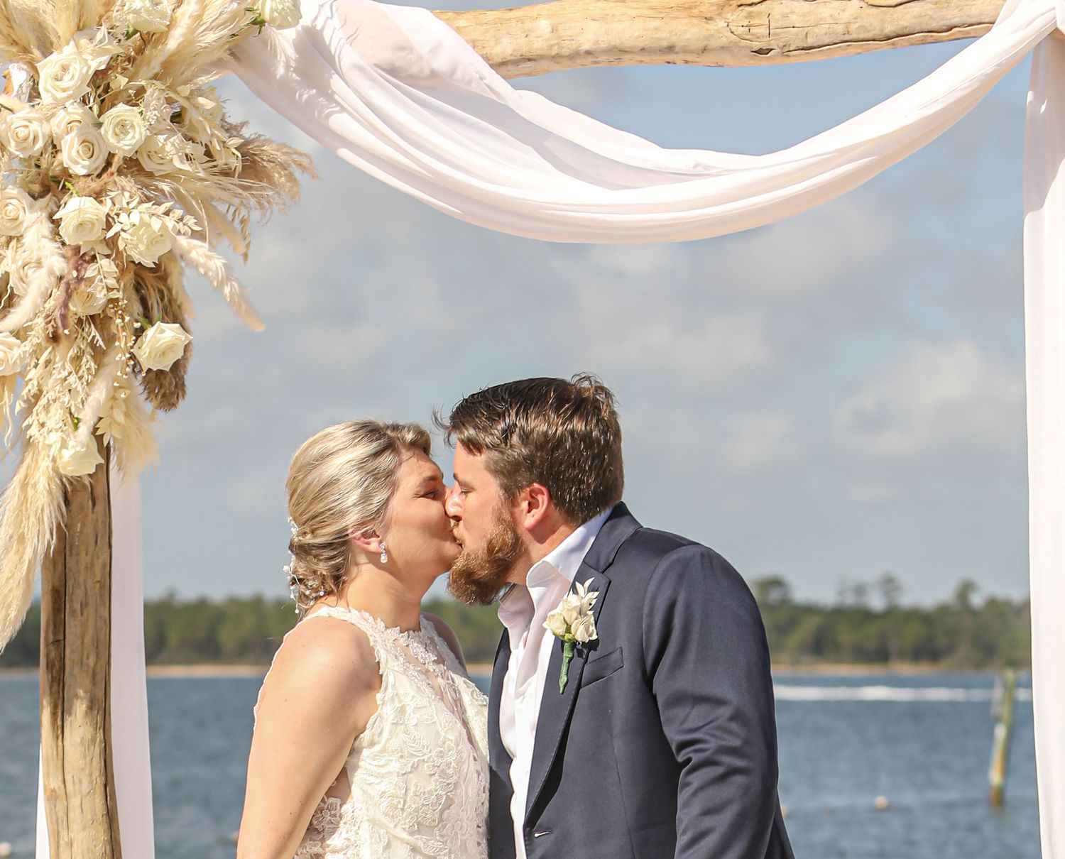 Wedding couple shares intimate moment under floral arch with water view.