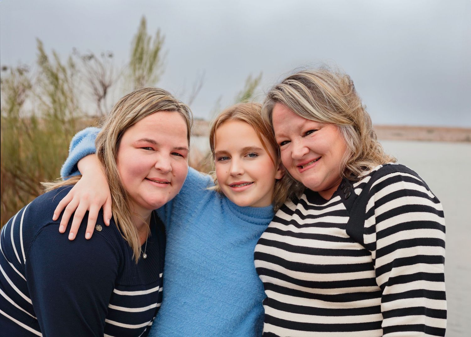 Three people pose together outdoors with arms around each other, wearing blue and striped tops against a scenic backdrop.