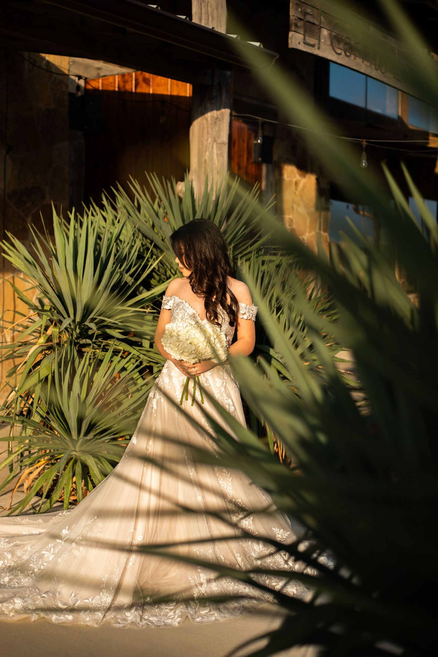A woman in a lace wedding dress poses in a garden setting with palm fronds in the foreground.
