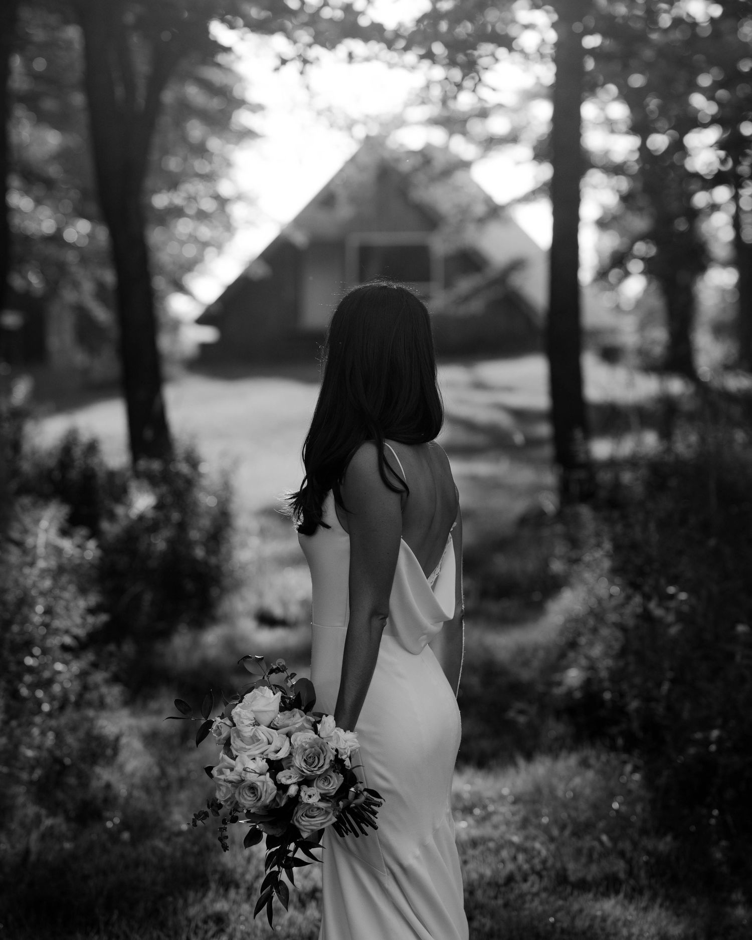 A silhouette of a bride holding a bouquet stands in a dreamy forest setting with a cabin in the background.