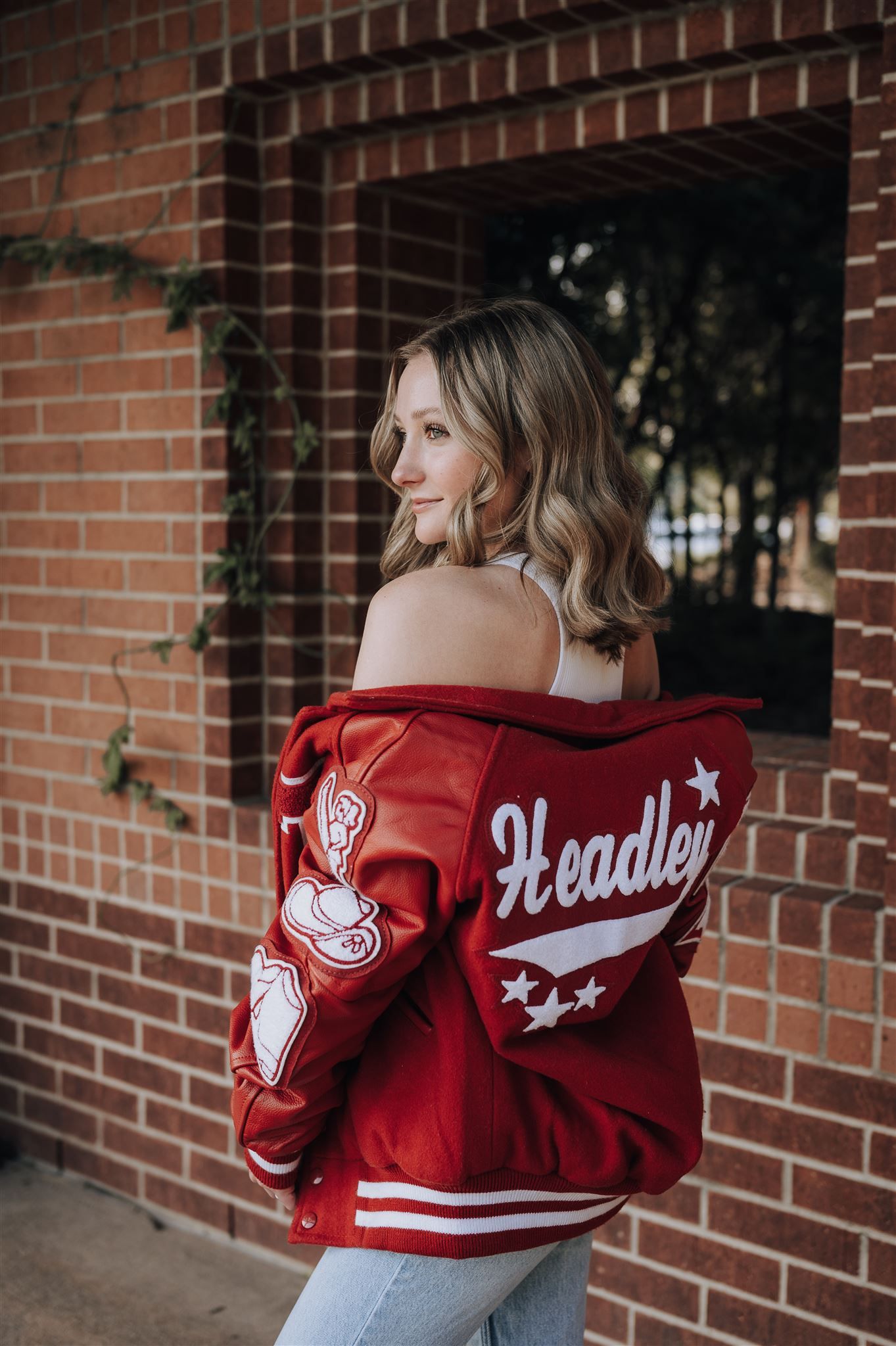 Side view of someone wearing a red varsity jacket with 'Headley' lettering against brick wall.